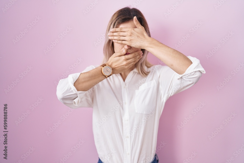 Young beautiful woman standing over pink background covering eyes and mouth with hands, surprised and shocked. hiding emotion Young beautiful woman standing over pink background covering eyes and mouth with hands, surprised and shocked. hiding emotion