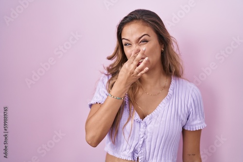 Young hispanic woman standing over pink background smelling something stinky and disgusting, intolerable smell, holding breath with fingers on nose. bad smell