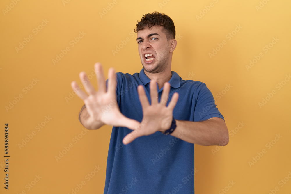 Young hispanic man standing over yellow background afraid and terrified with fear expression stop gesture with hands, shouting in shock. panic concept. Young hispanic man standing over yellow background afraid and terrified with fear expression stop gesture with hands, shouting in shock. panic concept.