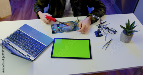 Top view. Unknown male hands using green screen tablet und soldering microcircuit with a tool repairs a home electronic appliance and screwdriwer on the table in evening at home. photo