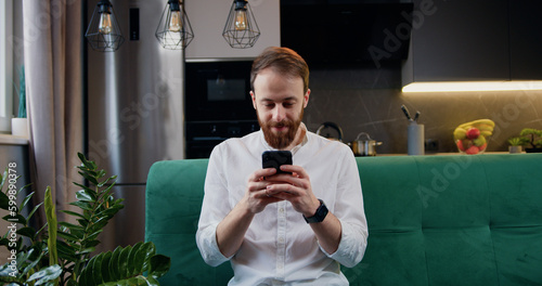 Handsome man browsing smartphone while sitting on sofa in kitchen at home. Cheerful bearded caucasian male in white shirt is texing and read a message. photo