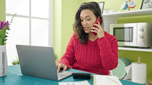 Young beautiful hispanic woman talking on smartphone accounting money at dinning room