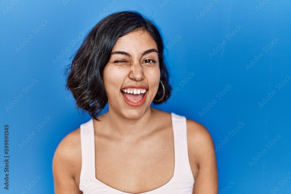 Young hispanic woman standing over blue background winking looking at the camera with sexy expression, cheerful and happy face. Young hispanic woman standing over blue background winking looking at the camera with sexy expression, cheerful and happy face.