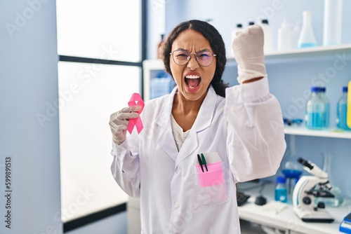 Young hispanic doctor woman working at scientist laboratory holding pink ribbon annoyed and frustrated shouting with anger  yelling crazy with anger and hand raised