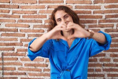 Beautiful brunette woman standing over bricks wall smiling in love doing heart symbol shape with hands. romantic concept.