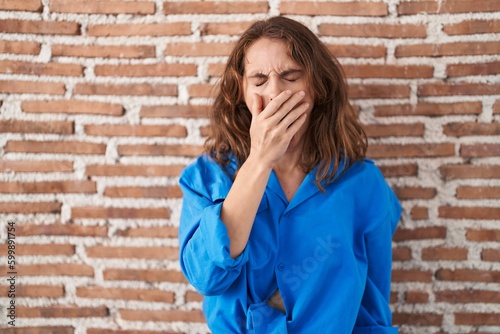 Beautiful brunette woman standing over bricks wall bored yawning tired covering mouth with hand. restless and sleepiness.