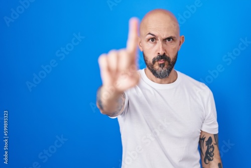 Hispanic man with tattoos standing over blue background pointing with finger up and angry expression, showing no gesture