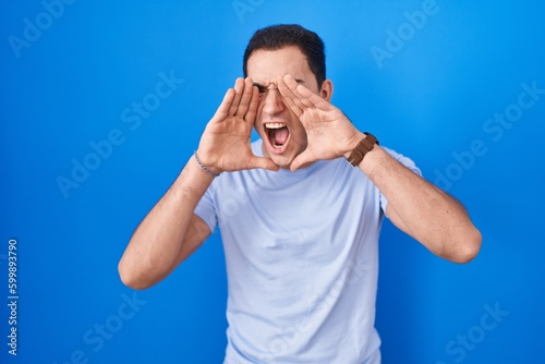 Young hispanic man standing over blue background shouting angry out loud with hands over mouth