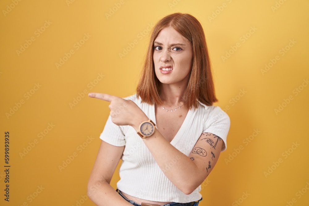 Young redhead woman standing over yellow background pointing aside worried and nervous with forefinger, concerned and surprised expression Young redhead woman standing over yellow background pointing aside worried and nervous with forefinger, concerned and surprised expression