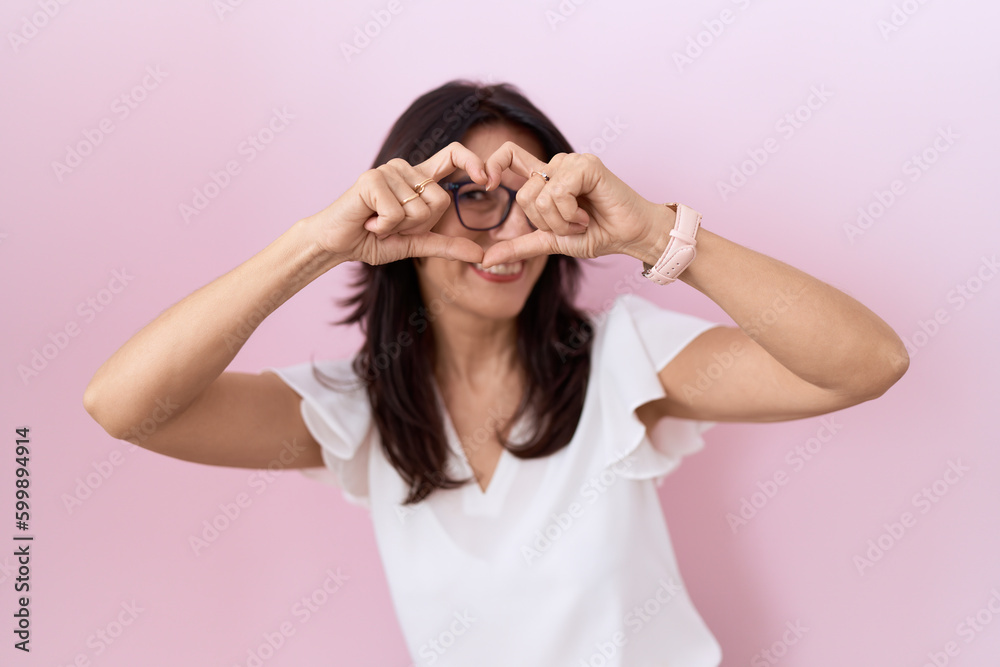 Middle age hispanic woman wearing casual white t shirt and glasses doing heart shape with hand and fingers smiling looking through sign Middle age hispanic woman wearing casual white t shirt and glasses doing heart shape with hand and fingers smiling looking through sign