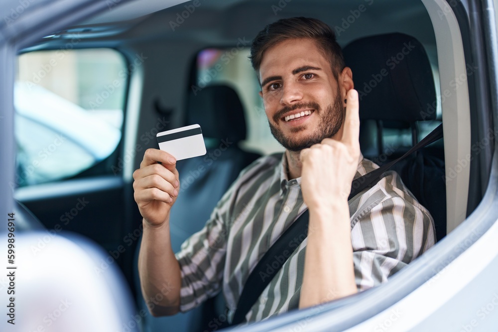 Hispanic man with beard driving car holding credit card smiling with an idea or question pointing finger with happy face, number one