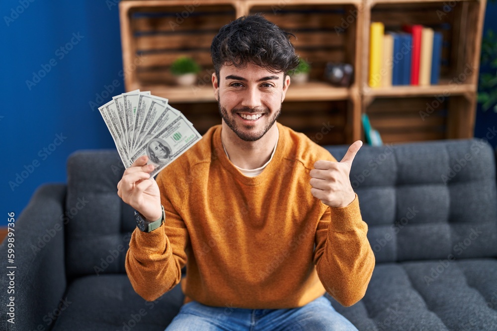 Hispanic man with beard holding 100 dollars banknotes smiling happy and positive, thumb up doing excellent and approval sign Hispanic man with beard holding 100 dollars banknotes smiling happy and positive, thumb up doing excellent and approval sign