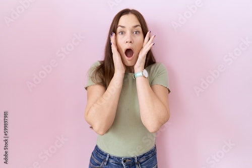 Beautiful brunette woman standing over pink background afraid and shocked, surprise and amazed expression with hands on face
