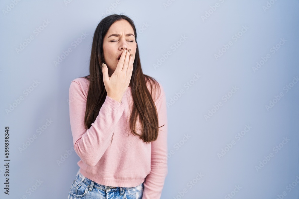 Young brunette woman standing over blue background bored yawning tired covering mouth with hand. restless and sleepiness. Young brunette woman standing over blue background bored yawning tired covering mouth with hand. restless and sleepiness.