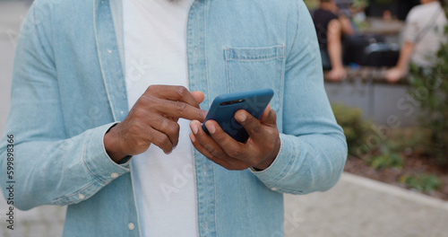 Unknown stylish black-skinned man in casual clothes standing outdoors near cafe and browsing phone apps,close up