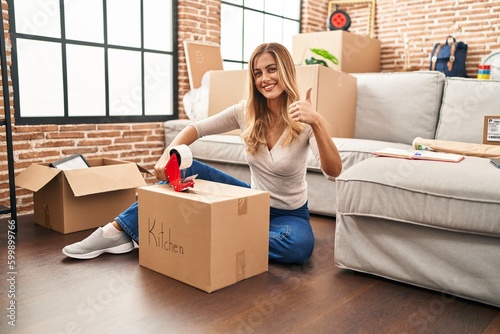 Young blonde woman sitting on the floor closing boxes home smiling happy and positive, thumb up doing excellent and approval sign © Krakenimages.com