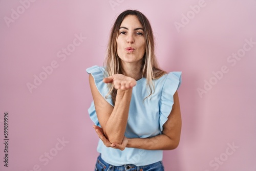 Young hispanic woman standing over pink background looking at the camera blowing a kiss with hand on air being lovely and sexy. love expression.
