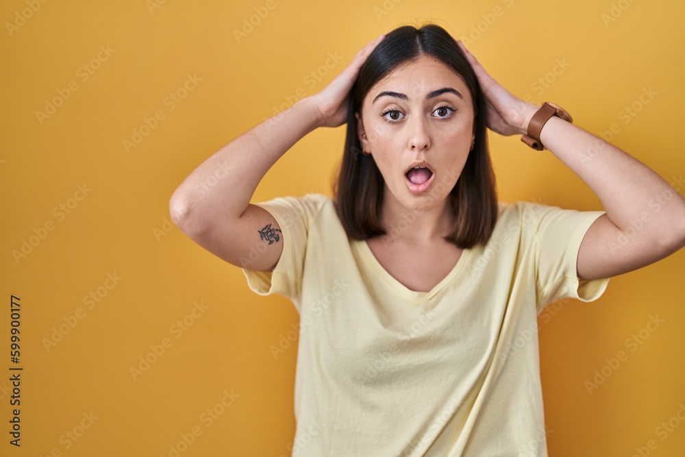 Hispanic girl wearing casual t shirt over yellow background crazy and scared with hands on head, afraid and surprised of shock with open mouth Hispanic girl wearing casual t shirt over yellow background crazy and scared with hands on head, afraid and surprised of shock with open mouth