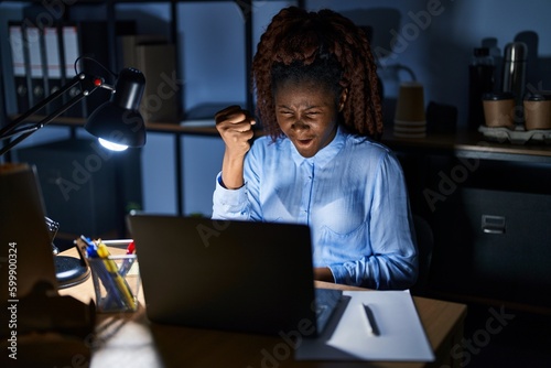 African woman working at the office at night angry and mad raising fist frustrated and furious while shouting with anger. rage and aggressive concept.