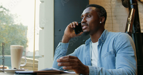 Attractive satisfeid smiling young african american sitting near the window of street cafe and enjoying his phone conversation at time break,close up