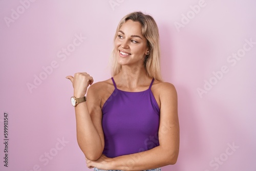 Young blonde woman standing over pink background smiling with happy face looking and pointing to the side with thumb up.