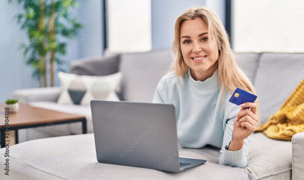 Young blonde woman using laptop and credit card lying on sofa at home Young blonde woman using laptop and credit card lying on sofa at home