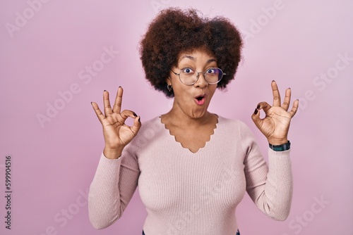 Young african american woman standing over pink background looking surprised and shocked doing ok approval symbol with fingers. crazy expression