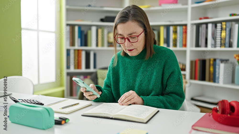 Mature hispanic woman with grey hair student using smartphone reading a book at library Mature hispanic woman with grey hair student using smartphone reading a book at library