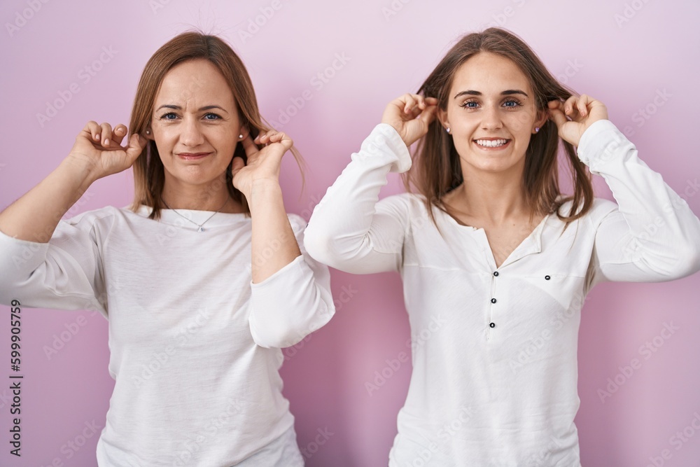 Middle age mother and young daughter standing over pink background smiling pulling ears with fingers, funny gesture. audition problem Middle age mother and young daughter standing over pink background smiling pulling ears with fingers, funny gesture. audition problem