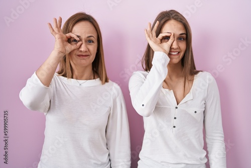Middle age mother and young daughter standing over pink background doing ok gesture with hand smiling, eye looking through fingers with happy face.