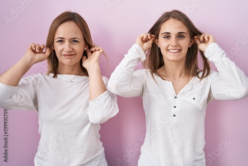Middle age mother and young daughter standing over pink background smiling pulling ears with fingers, funny gesture. audition problem