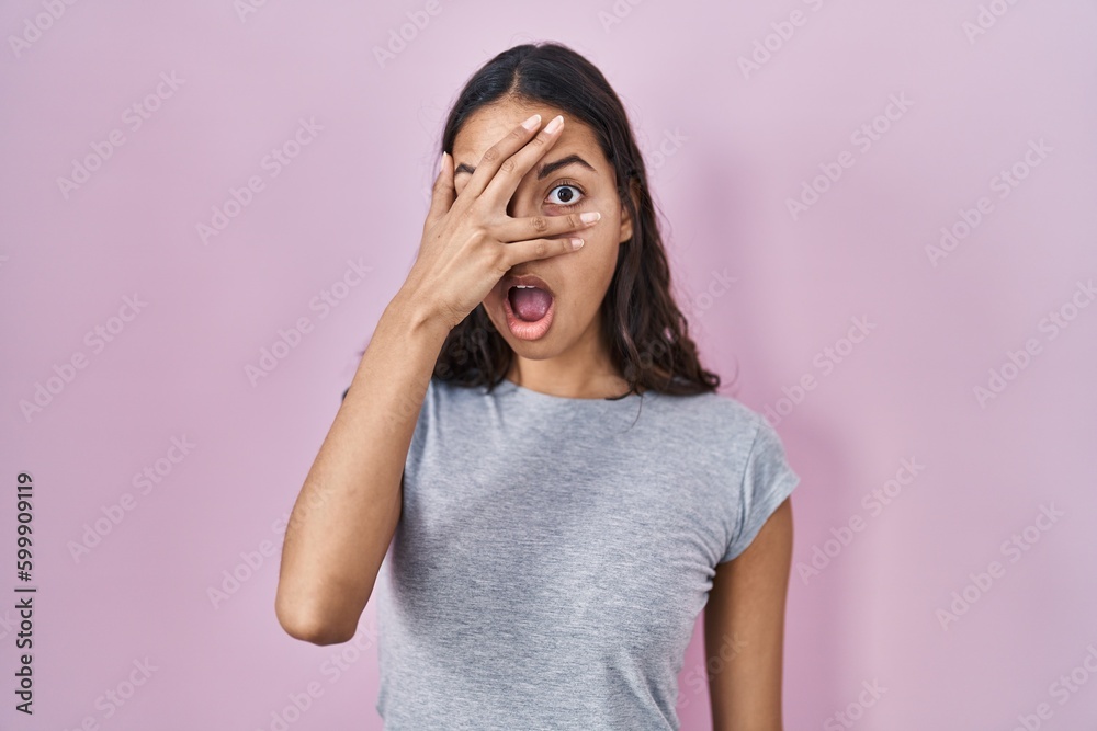 Young brazilian woman wearing casual t shirt over pink background peeking in shock covering face and eyes with hand, looking through fingers with embarrassed expression. Young brazilian woman wearing casual t shirt over pink background peeking in shock covering face and eyes with hand, looking through fingers with embarrassed expression.