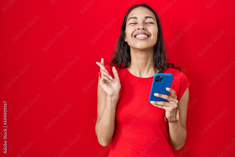 Young brazilian woman using smartphone over red background gesturing finger crossed smiling with hope and eyes closed. luck and superstitious concept. Young brazilian woman using smartphone over red background gesturing finger crossed smiling with hope and eyes closed. luck and superstitious concept.