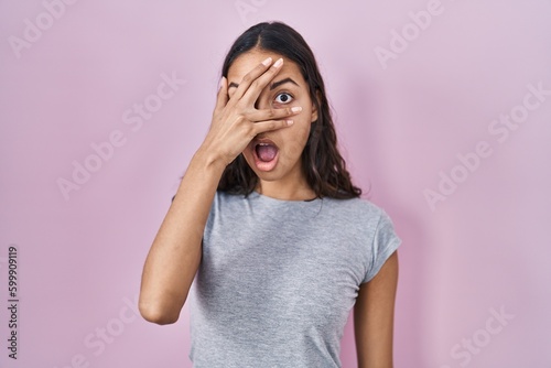 Young brazilian woman wearing casual t shirt over pink background peeking in shock covering face and eyes with hand, looking through fingers with embarrassed expression. © Krakenimages.com
