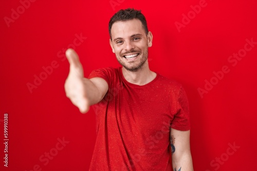 Young hispanic man standing over red background smiling friendly offering handshake as greeting and welcoming. successful business.