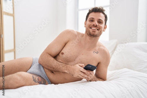 Young hispanic man using smartphone lying on bed at bedroom