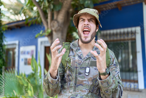 Young hispanic man wearing camouflage army uniform outdoors crazy and mad shouting and yelling with aggressive expression and arms raised. frustration concept.