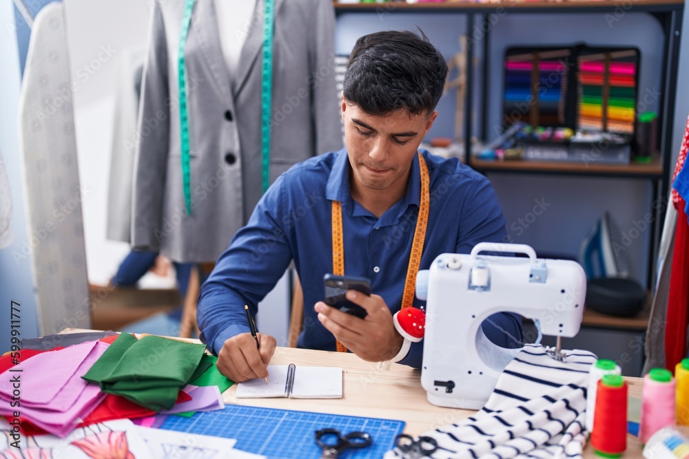 Young hispanic man tailor using smartphone writing on notebook at clothing factory