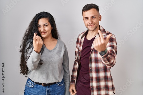 Young hispanic couple standing over white background beckoning come here gesture with hand inviting welcoming happy and smiling