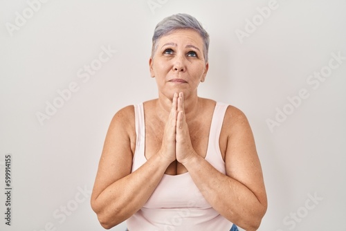 Middle age caucasian woman standing over white background praying with hands together asking for forgiveness smiling confident.
