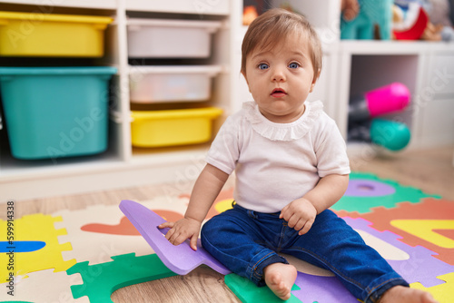 Adorable hispanic baby sitting on floor with relaxed expression at kindergarten