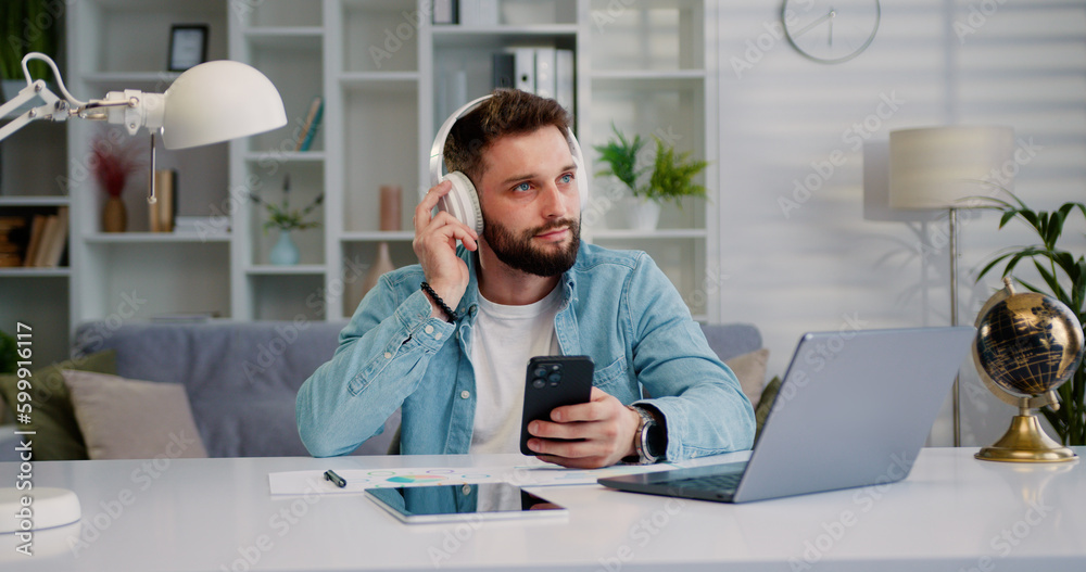 Modern, happy and relaxed man with headphones holding mobile phone watching videos and listening to favorite music podcast or online social media at home office. Modern, happy and relaxed man with headphones holding mobile phone watching videos and listening to favorite music podcast or online social media at home office.