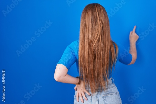 Redhead woman standing over blue background posing backwards pointing ahead with finger hand