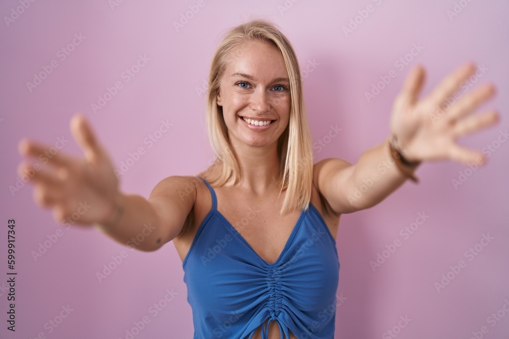 Fototapeta premium Young caucasian woman standing over pink background looking at the camera smiling with open arms for hug. cheerful expression embracing happiness.
