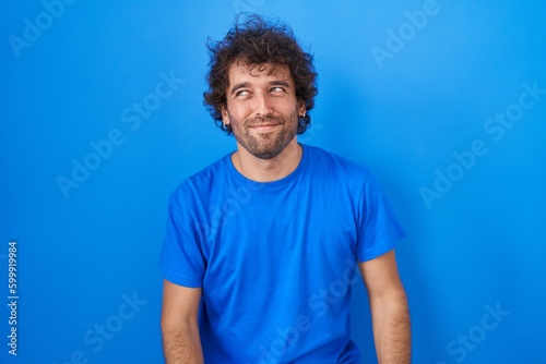 Hispanic young man standing over blue background smiling looking to the side and staring away thinking.