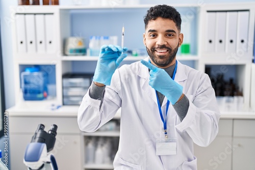 Hispanic man with beard working at scientist laboratory holding syringe smiling happy pointing with hand and finger