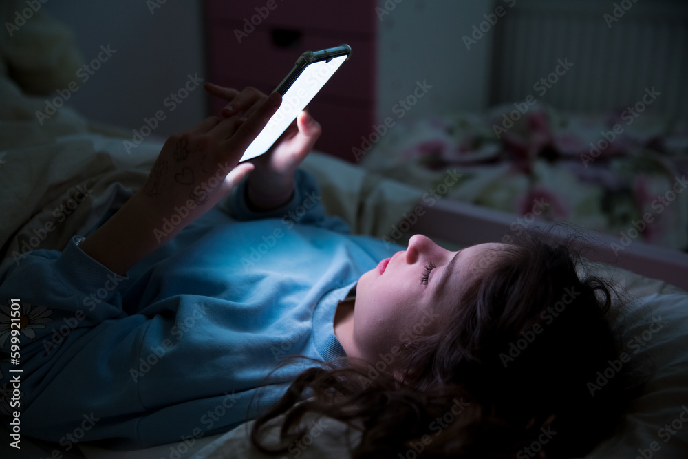 A child using smart phone lying in bed late at night, playing games, watching videos online, scrolling screen. Children's screen addiction and parent control concept. Child's room at night.  - obrazy, fototapety, plakaty 