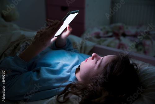 Fototapeta A child using smart phone lying in bed late at night, playing games, watching videos online, scrolling screen