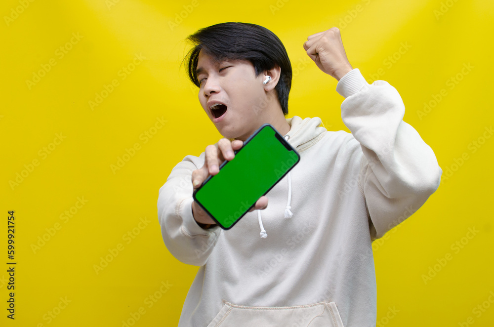 Asian man in beige hoodie using earphones, raising fist and showing his screen phone with green screen template best for advertisment. man showing mock up of phone. Asian man in beige hoodie using earphones, raising fist and showing his screen phone with green screen template best for advertisment. man showing mock up of phone.