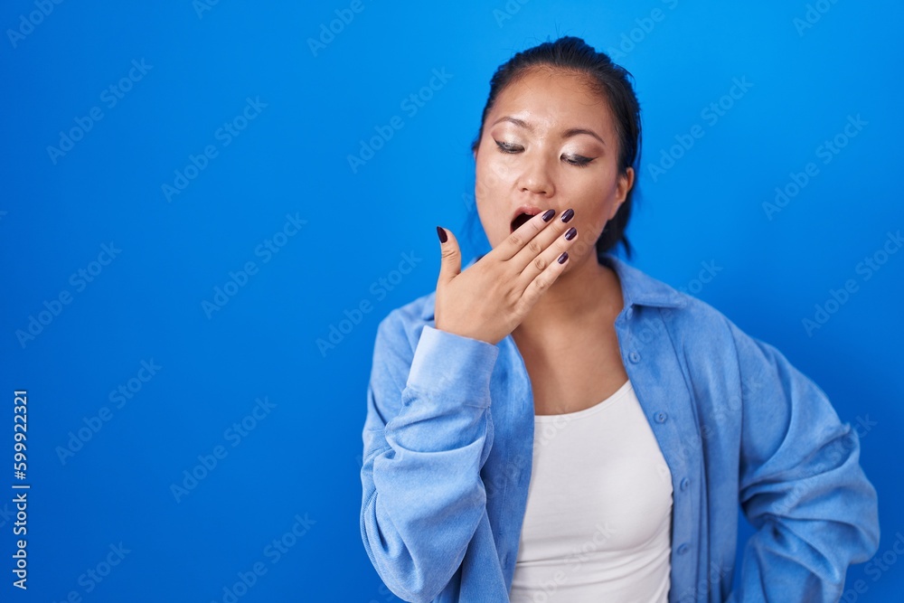 Asian young woman standing over blue background bored yawning tired covering mouth with hand. restless and sleepiness. Asian young woman standing over blue background bored yawning tired covering mouth with hand. restless and sleepiness.